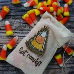 A muslin drawstring bag with a burlap candy-corn patch and “Got Candy?” text, surrounded by candy corn pieces, on a rustic wooden surface.