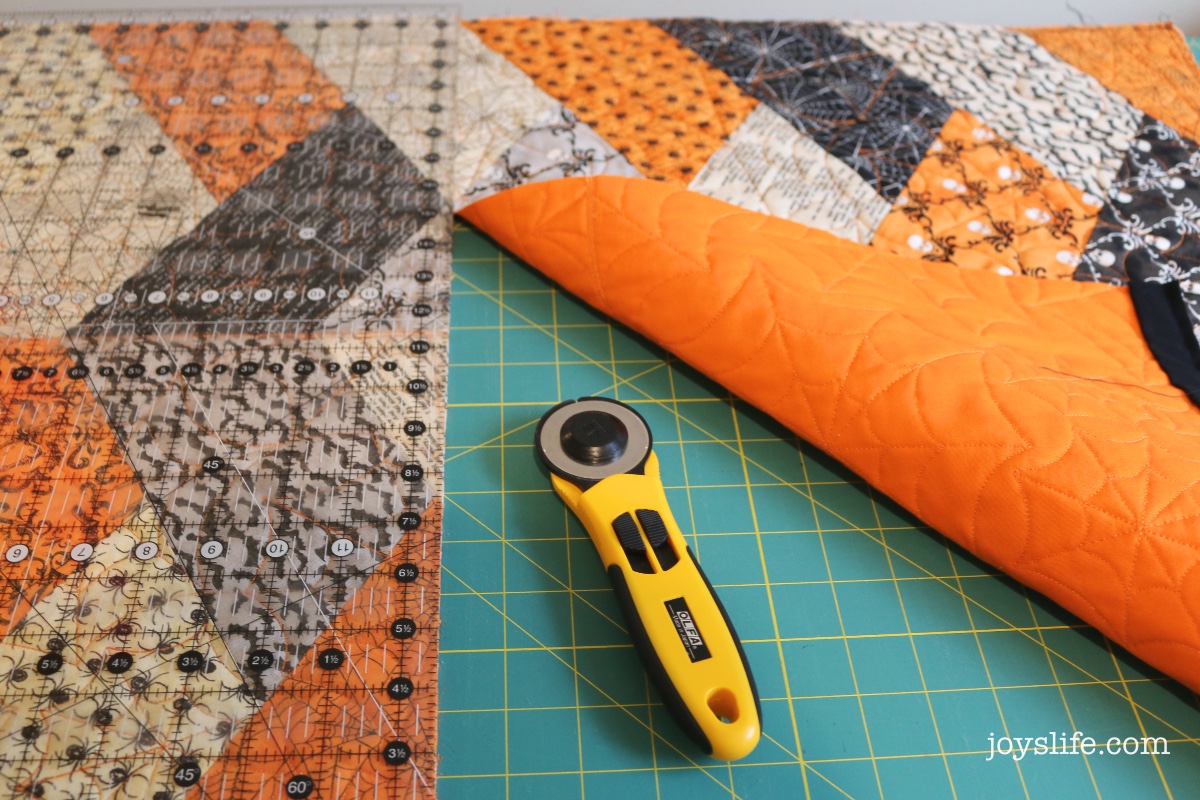Quilted Halloween fabric on a green cutting mat with clear quilting ruler and yellow Olfa rotary cutter, showing orange quilted backing and stitched design.