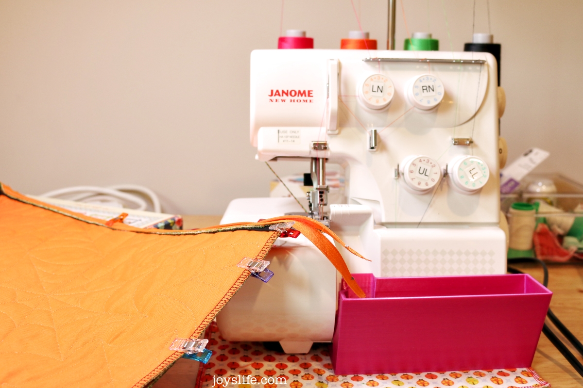 Janome serger sewing orange quilted pillowcase fabric with clips holding edges in place and a pink bin catching fabric trimmings.