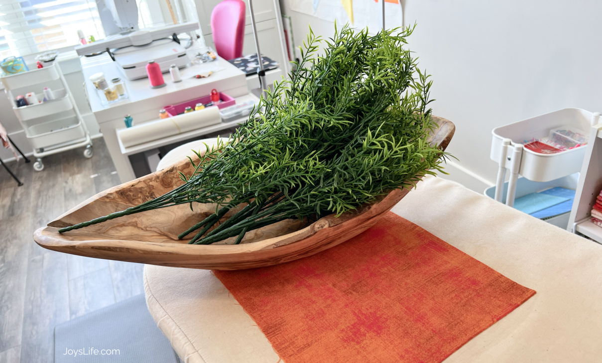 Artificial carrot stems arranged in a wooden bowl on a sewing room table for a fabric Easter carrot project tutorial at JoysLife.com where you can learn to sew this beginner friendly decoration