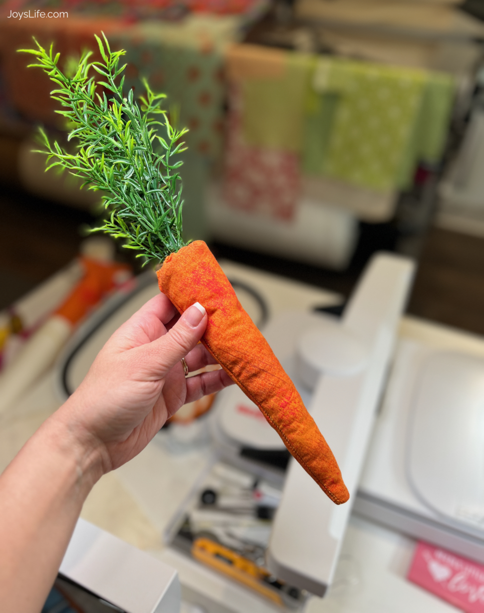 Hand holding a finished fabric Easter carrot with faux greenery stem top in a sewing room workspace, tutorial at joyslife.com