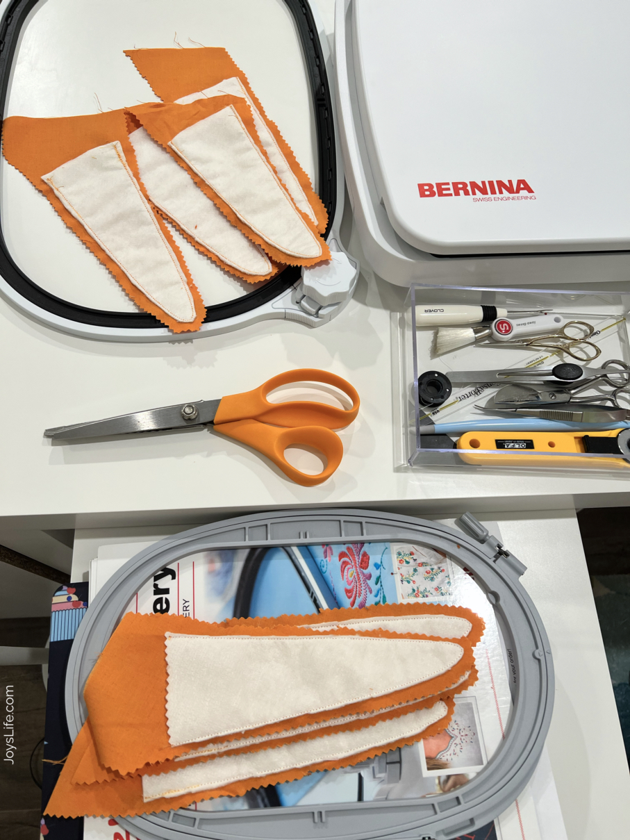 Fabric Easter carrot pieces with batting and orange fabric arranged near an embroidery hoop, scissors, and sewing tools in a sewing workspace, tutorial at joyslife.com