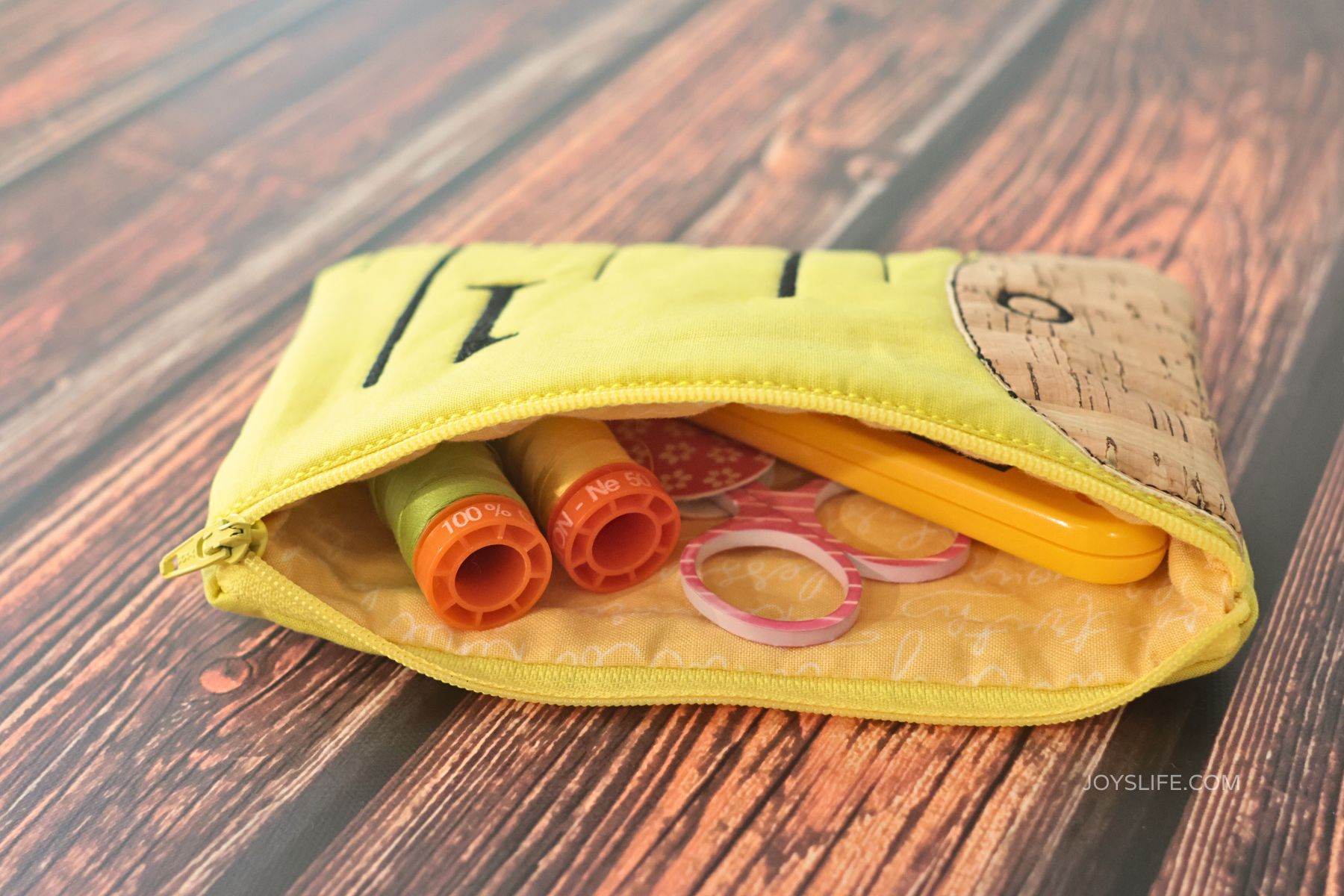 Open yellow Tape Measure Clutch embroidery bag with cork fabric end, filled with thread spools, clips, and sewing notions on a wooden background.