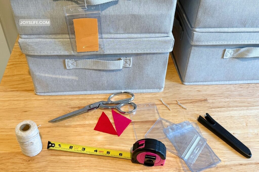 Fabric scrap storage tools on a table: gray bins, clear tag holders, scissors, tape measure, twine, and fabric swatches for making color coded storage.