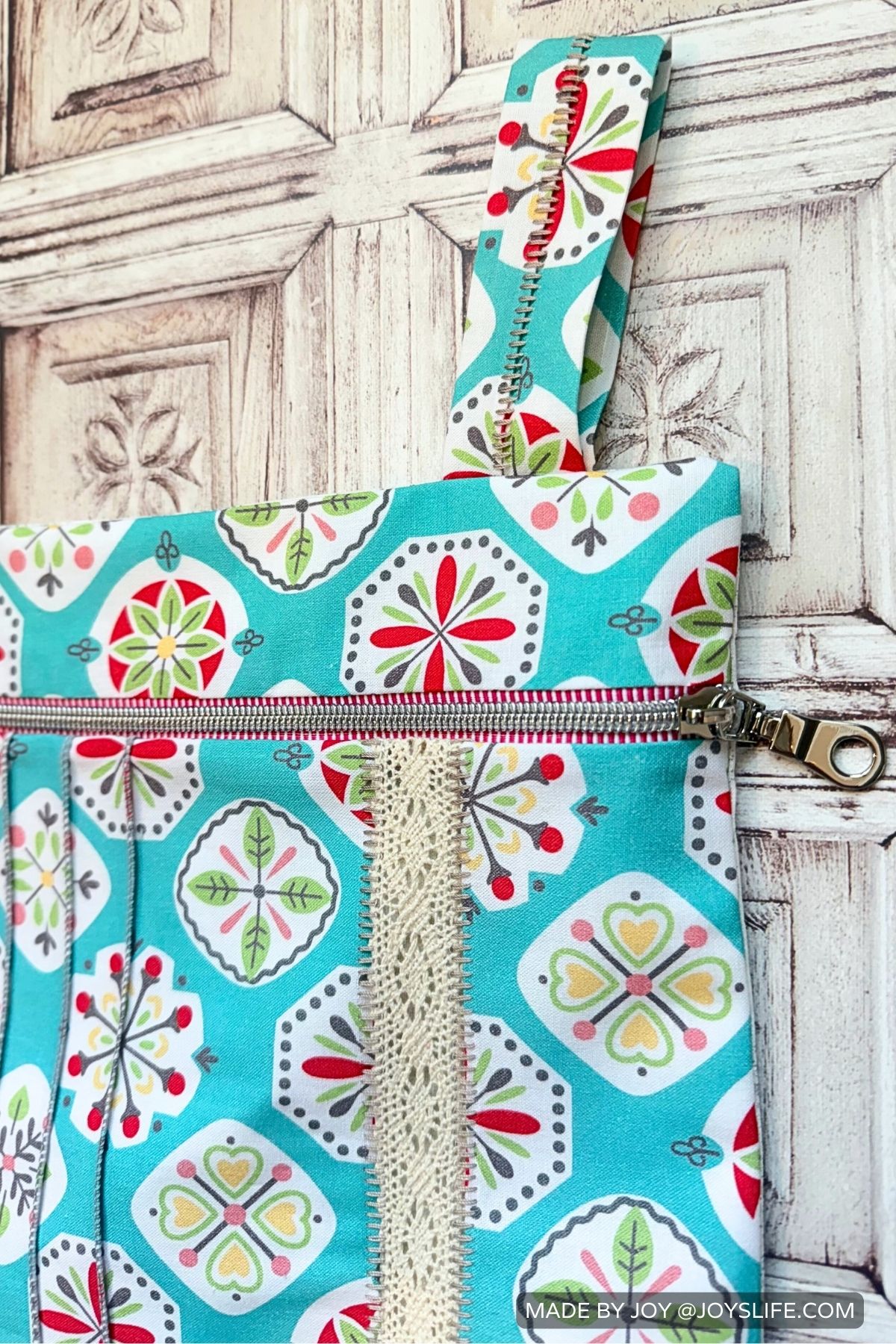 Close-up of the Hideaway Hanger Safe sewing project showing a red and silver zipper, lace insertion, and fabric strap. The teal geometric floral fabric with red, green, and yellow accents is stitched with serger seams and displayed against a rustic white wooden background.