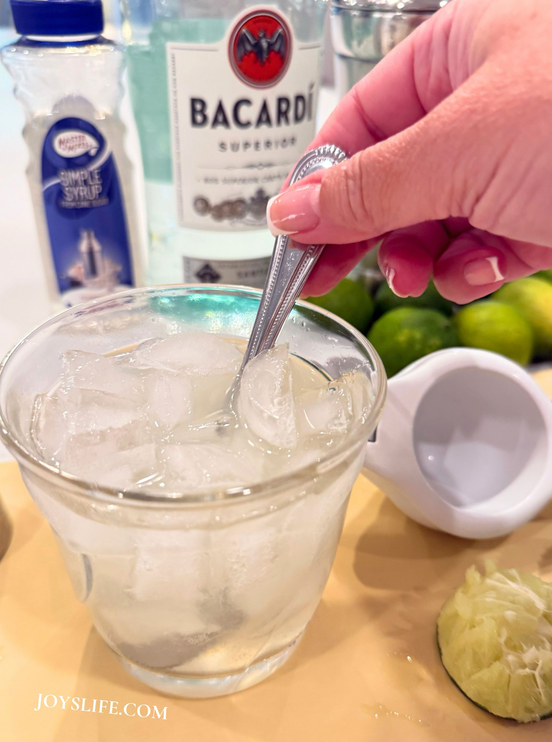 Hand stirring a daiquiri in a rocks glass with ice, Bacardi rum, simple syrup, limes, and a citrus press in the background.