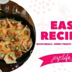 Cast iron skillet filled with cheesy nachos topped with sliced eggrolls, and hot sauce, placed on a blue tea towel with silverware.