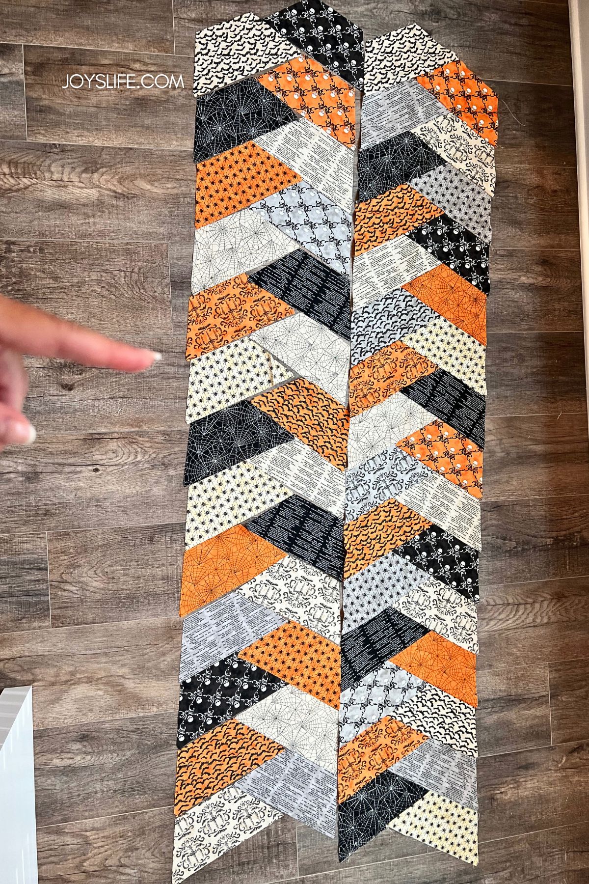 Halloween quilted table runner layout on wood floor with orange, black, and cream fabrics featuring bats, spiders, spiderwebs, skulls, and text prints.