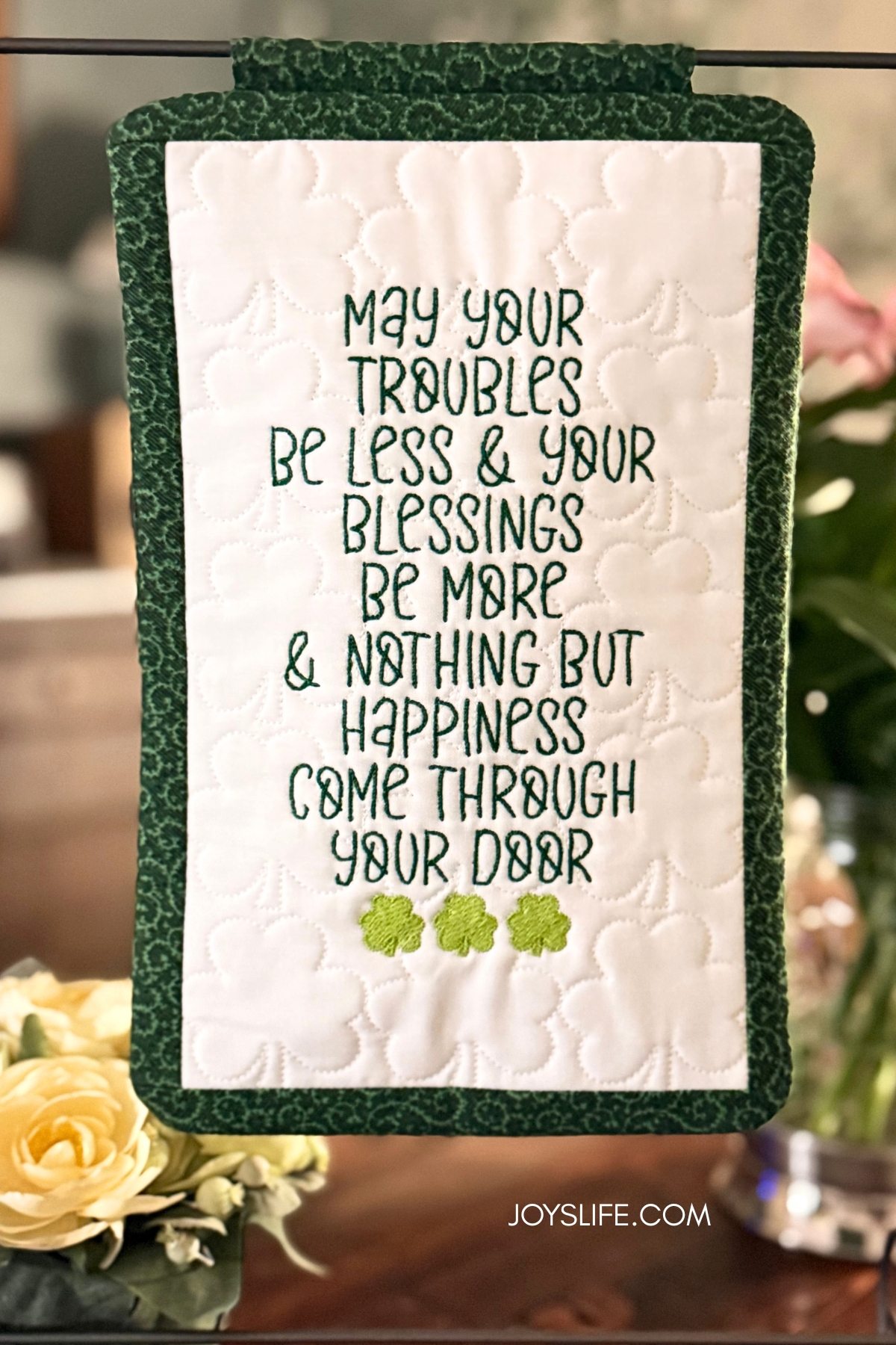 Alt Text Irish blessing mini quilt with shamrock quilting and embroidered lettering displayed on a stand. Find the tutorial at JoysLife.com.