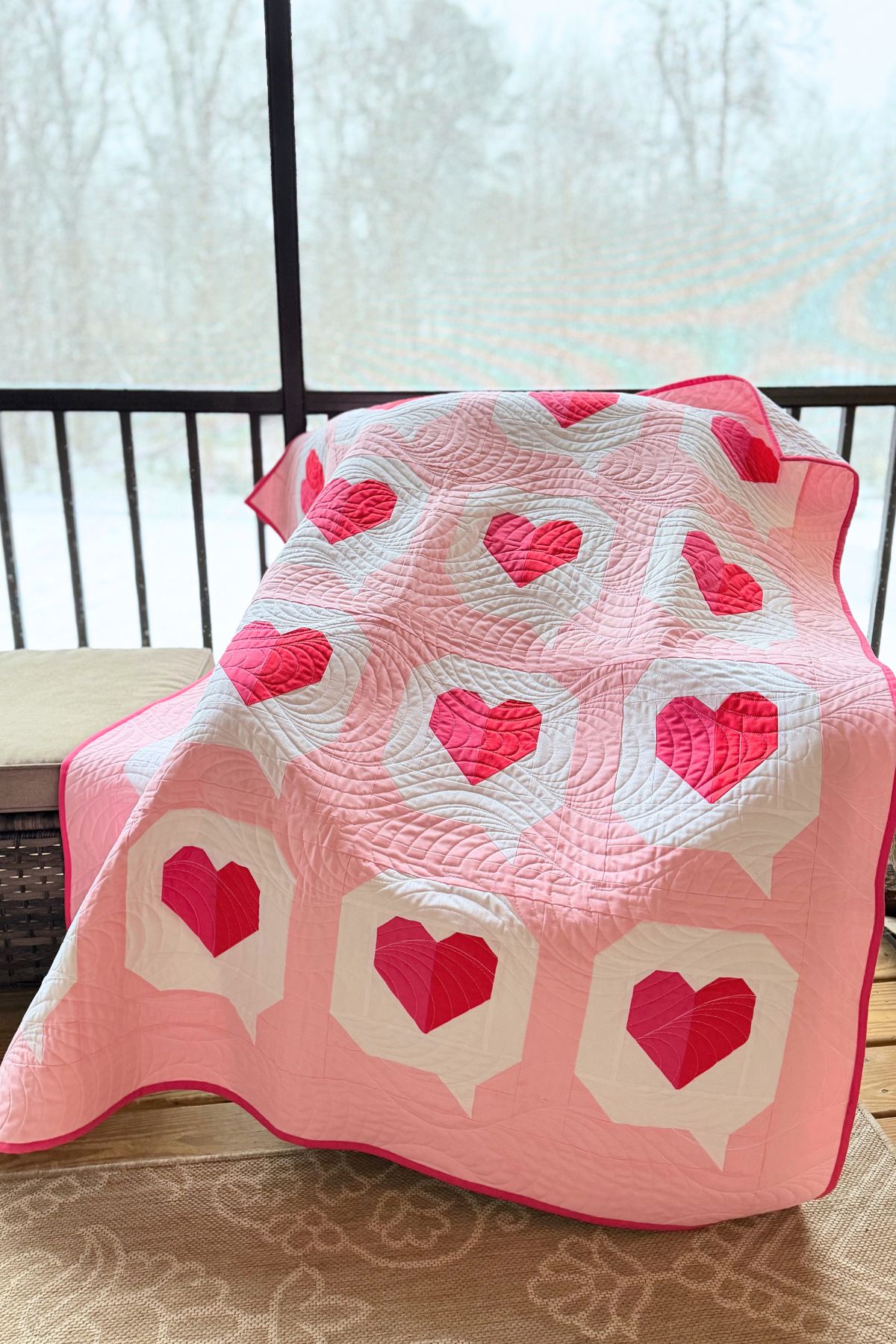 Heart quilt with pink and red heart pattern, longarm quilted laying over a chair in a covered porch with snow falling outside