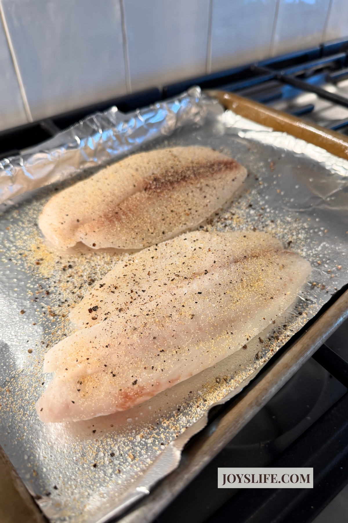 Raw tilapia fillets seasoned with garlic powder on a foil lined baking sheet before baking for tilapia fish tacos on joyslife.com